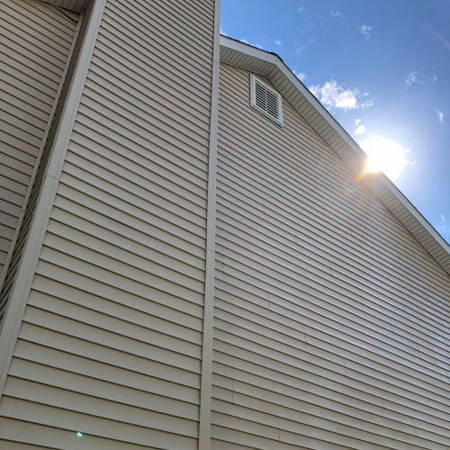 A tall, beige building with vertical siding is shown from a low angle. The sun is partially visible on the right side of the image, casting a bright flare against the clear blue sky with a few scattered clouds. This structure stands as a testament to impeccable St. Charles remodeling craftsmanship.